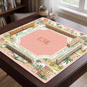 Puede incluir: Un juego de Mahjong sobre una mesa. El tablero presenta un diseño tropical rosa y verde con un monograma central. Las fichas de madera están dispuestas alrededor del tablero, listas para jugar.