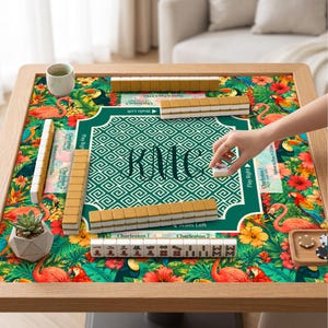 May include: A Mahjong game set on a square table with a tropical print featuring flamingos, toucans, and hibiscus flowers. The center mat has a green geometric pattern with the monogram KMC. Includes ivory-colored tiles and a small succulent plant.