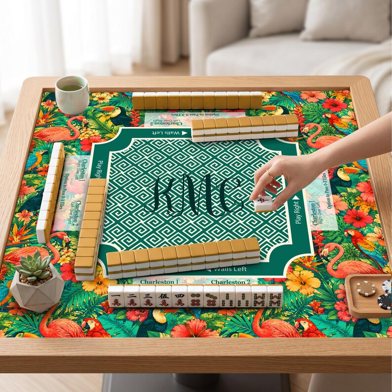 May include: A Mahjong game set on a square table with a tropical print featuring flamingos, toucans, and hibiscus flowers. The center mat has a green geometric pattern with the monogram KMC. Includes ivory-colored tiles and a small succulent plant.