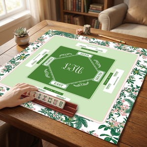 Pode incluir: Um tapete de jogo de Mahjong verde e branco com uma borda floral. O centro do tapete é verde com as palavras "Norte", "Sul", "Este" e "Oeste" em branco. Um suporte de azulejos de madeira com azulejos está em primeiro plano.
