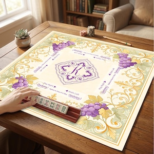 May include: A square Mahjong mat featuring a cream-colored center with a purple letter K monogram, surrounded by a decorative border of purple grapes and green leaves. The mat includes printed game instructions and a wooden tile rack with tiles.