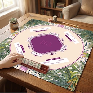 以下が含まれることがあります： 花柄とヤシの葉のデザインの麻雀ゲームセット。ゲーム盤はベージュで、紫色の中心があり、「北」、「東」、「南」、「西」の文字があります。手は、タイルが入った木製のタイルホルダーを持っています。