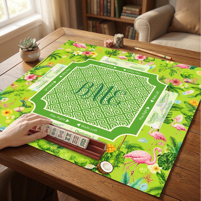 K&ouml;nnte beinhalten: Eine quadratische Mahjong-Matte mit einem leuchtend gr&uuml;nen Hintergrund und einem tropischen Muster aus rosa Flamingos, Hibiskusbl&uuml;ten und Palmenbl&auml;ttern. Die Mitte zeigt einen wei&szlig;en geometrischen Rand mit dem Monogramm BMc und Spielanleitungen.