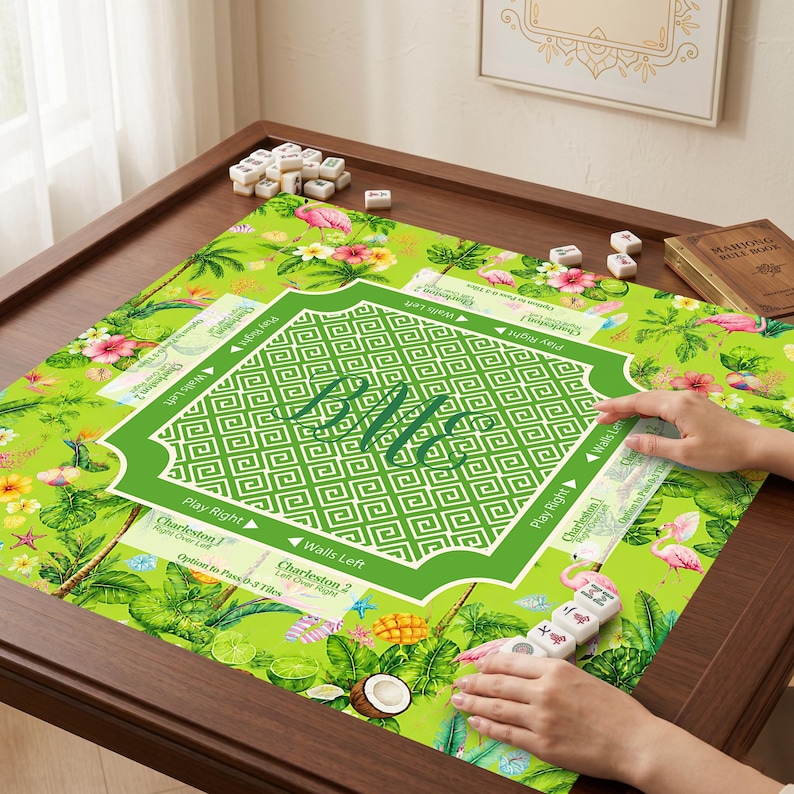 K&ouml;nnte beinhalten: Eine quadratische Mahjong-Matte mit einem leuchtend gr&uuml;nen Tropenmuster, rosa Flamingos, Hibiskusbl&uuml;ten und Palmenbl&auml;ttern. Die Mitte hat einen wei&szlig;en geometrischen Rand mit dem Monogramm BLC. Der Text umfasst Charleston 1, Charleston 2, Play Right und Walls Left.