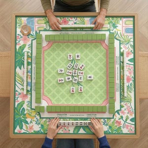 May include: A top-down view of a Mahjong game in progress. The game board is a light wood frame with a floral and botanical design. Green and white tiles are arranged on the board, with some tiles scattered in the center. The text "CHARLESTON" is visible.