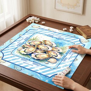 May include: A Mahjong game set with a blue and white oyster-themed game board. The board features illustrations of oysters, seashells, and aquatic elements. The game board is on a dark brown wooden table, with Mahjong tiles and a rule book.
