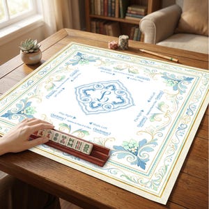 May include: A square Mahjong mat with a white center featuring a blue monogram L, surrounded by blue and green floral scrollwork. The mat includes printed game instructions like Charleston and Walls Left. A wooden rack holds five Mahjong tiles.