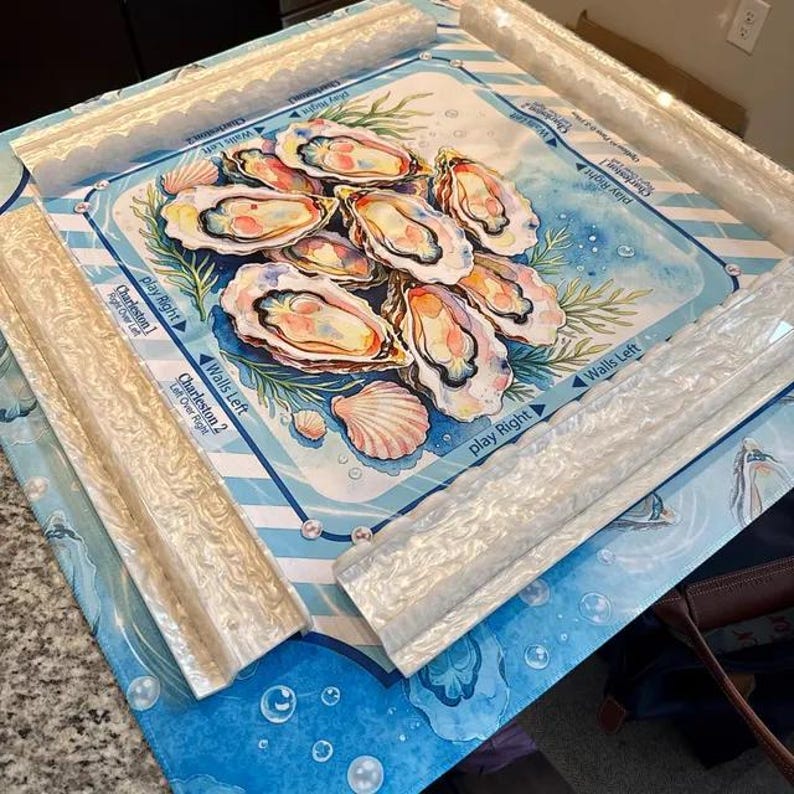 May include: A square mahjong mat featuring a central watercolor illustration of oysters and seashells on a blue background. The mat includes white text labels such as Charleston 1, Charleston 2, Walls Left, and Play Right, framed by four white textured racks.