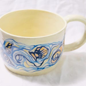 May include: Cream-colored ceramic mug with a large handle. The mug features a hand-painted design of blue and yellow waves with a seashell motif. The interior of the mug is a pale yellow color.
