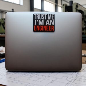 May include: A silver laptop with a sticker that reads "TRUST ME I'M AN ENGINEER" in white and red text on a black background. The laptop is on a surface with architectural plans.