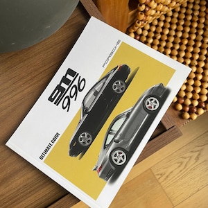 May include: A white book titled "911 996 ULTIMATE GUIDE" with two Porsche 911 cars on a yellow background. The book is open on a wooden surface, with a wooden beaded chair in the background.