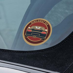 May include: A circular sticker featuring the text "NINE EIGHT SIX" around a car illustration. The car is navy blue with red lights, set against a maroon background with a gold border. The sticker is attached to a car window.
