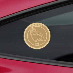 May include: A gold-coloured circular sticker with the word "STUTTGART" in a ring around a central design. The design includes a stylized shape and the number "996" at the bottom. The sticker is affixed to a car window.