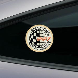 May include: A circular sticker featuring a globe graphic with a black and white checkered pattern on the left side. The text NINE NINE SEVEN and FLAT SIX ARCHIVE surrounds the globe, with the red number 997 centred across the middle of the design.