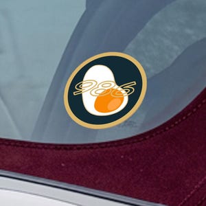 Puede incluir: Adhesivo circular con un diseño de huevo estilizado y el número "985" en blanco. El huevo tiene una parte superior blanca y una yema naranja, sobre un fondo azul marino con borde dorado. Para coche o ventana.