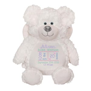 Puede incluir: Un oso de peluche blanco con detalles bordados. El oso tiene una cara sonriente, ojos azules y una nariz plateada. El pecho del oso presenta texto bordado con un nombre, un peso de 3,6 kg, una altura de 50,8 cm, junto con una fecha y hora.