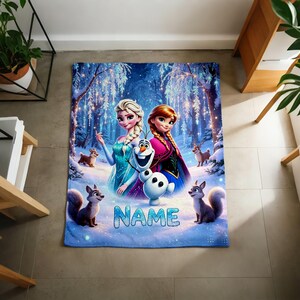 Puede incluir: Una manta rectangular de forro polar con una vibrante escena de bosque nevado con Elsa, Anna y Olaf. El dise&ntilde;o incluye peque&ntilde;os animales del bosque y la palabra NAME en letras grandes de color azul claro en la parte inferior del estampado.
