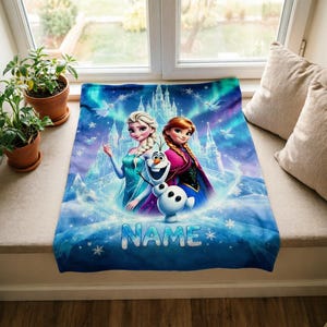 Puede incluir: Una manta rectangular de forro polar con una vibrante escena invernal en azul y morado con Elsa, Anna y Olaf de Frozen. El fondo muestra un castillo de hielo y copos de nieve. La palabra NAME est&aacute; impresa en letras grandes de color azul claro.
