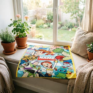May include: A rectangular blanket featuring a colorful print of various animated characters from the Toy Story franchise, including Woody, Buzz Lightyear, and Jessie, with a central red rectangular banner containing the white text NAME.