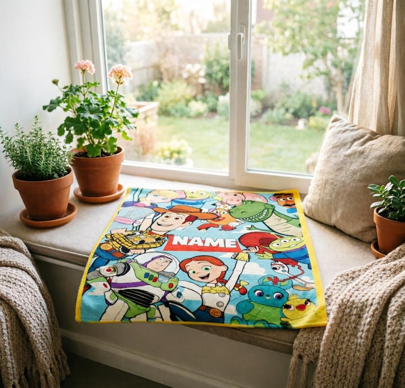 May include: A rectangular blanket featuring a colorful print of various animated characters from the Toy Story franchise, including Woody, Buzz Lightyear, and Jessie, with a central red rectangular banner containing the white text NAME.