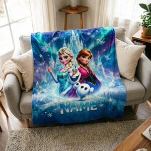 Puede incluir: Una manta de forro polar suave con una vibrante escena azul y morada con Elsa, Anna y Olaf de Frozen. El fondo muestra un castillo helado y copos de nieve. La palabra NAME est&aacute; impresa en letras grandes de color azul claro en el centro.