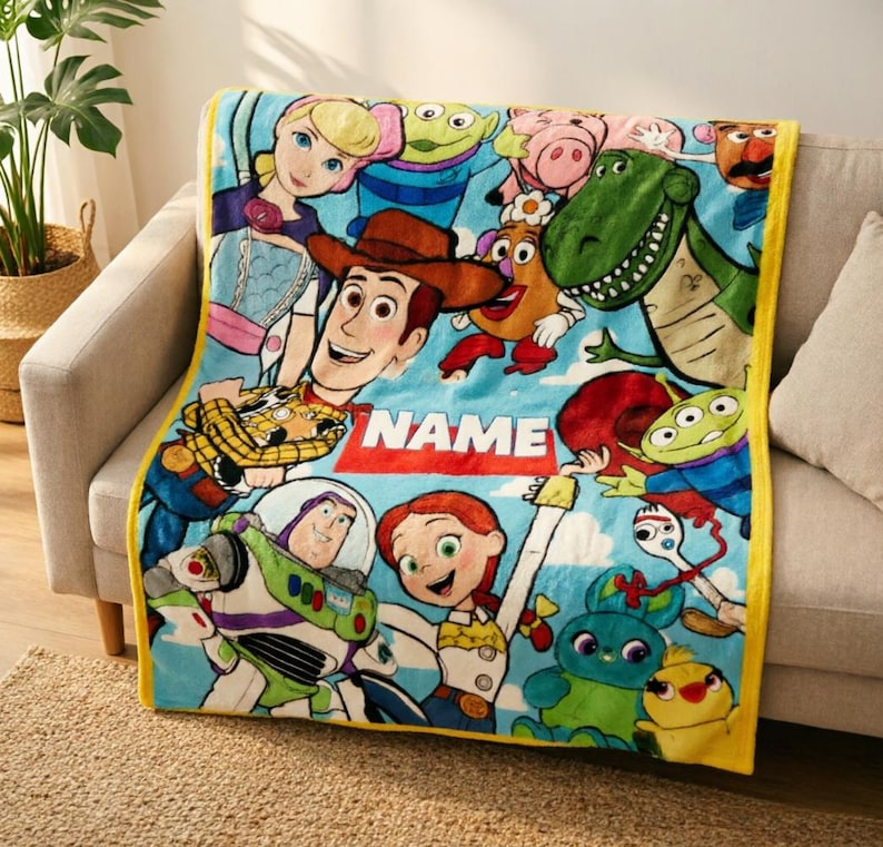 May include: A soft blue fleece throw blanket featuring a colorful print of Toy Story characters including Woody, Buzz Lightyear, Jessie, and Rex. The blanket has a bright yellow border and a central red rectangular area with the word NAME.