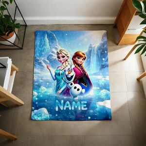 Puede incluir: Una manta rectangular de forro polar con un estampado gr&aacute;fico colorido de Elsa, Anna y Olaf de Frozen sobre un paisaje de monta&ntilde;a nevado con una cascada. La palabra NAME est&aacute; impresa en letras grandes, blancas y texturizadas en la parte inferior.