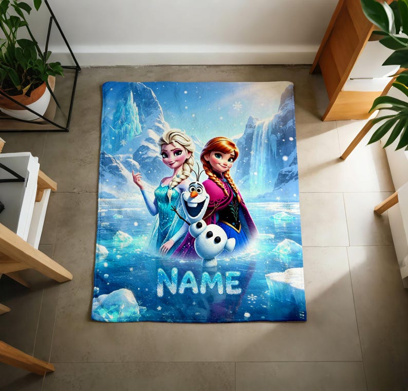 Puede incluir: Una manta rectangular de forro polar con un estampado gr&aacute;fico colorido de Elsa, Anna y Olaf de Frozen sobre un paisaje de monta&ntilde;a nevado con una cascada. La palabra NAME est&aacute; impresa en letras grandes, blancas y texturizadas en la parte inferior.