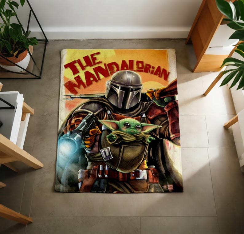 Może przedstawiać: Prostokątny dywan z grafiką The Mandalorian i Grogu z serii Gwiezdne Wojny. Dywan ma ż&oacute;łto-pomarańczowe tło z czerwonym napisem "The Mandalorian". Mandalorian jest przedstawiony w zbroi, trzymając broń i Grogu.