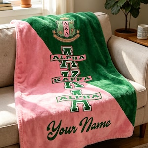 Op de afbeelding: Een roze en groene pluche deken met de Griekse letters Alpha Kappa Alpha in groen. De deken heeft een diagonaal kleurblokontwerp. De woorden "Your Name" staan in groene letters onderaan.