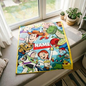 May include: A rectangular fleece blanket featuring a colorful print of various Toy Story characters including Woody, Buzz Lightyear, Jessie, and Rex on a light blue background. A prominent red rectangular patch displays the white text NAME.