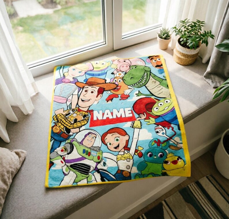 May include: A rectangular fleece blanket featuring a colorful print of various Toy Story characters including Woody, Buzz Lightyear, Jessie, and Rex on a light blue background. A prominent red rectangular patch displays the white text NAME.
