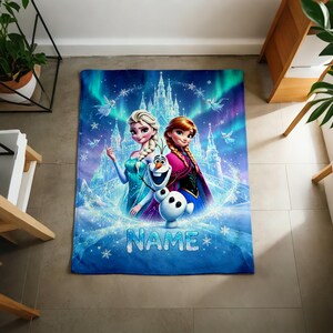 Puede incluir: Una manta rectangular de forro polar con un estampado gr&aacute;fico azul y morado de Elsa, Anna y Olaf de Frozen frente a un castillo de hielo. El dise&ntilde;o incluye copos de nieve, efectos de aurora boreal y la palabra NAME en texto azul.