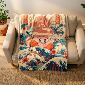 May include: A cream-colored throw blanket featuring a graphic print of a pink Kirby character surrounded by stylized blue ocean waves, pink cherry blossoms, and a soft pink sunset sky, with two vertical red rectangular panels containing text.