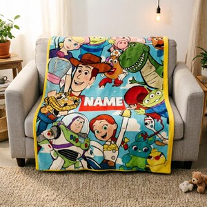 May include: A soft fleece throw blanket featuring a colorful Toy Story character print with Woody, Buzz Lightyear, Jessie, and other figures on a light blue background. The blanket has a yellow border and a central white rectangle with the text NAME.