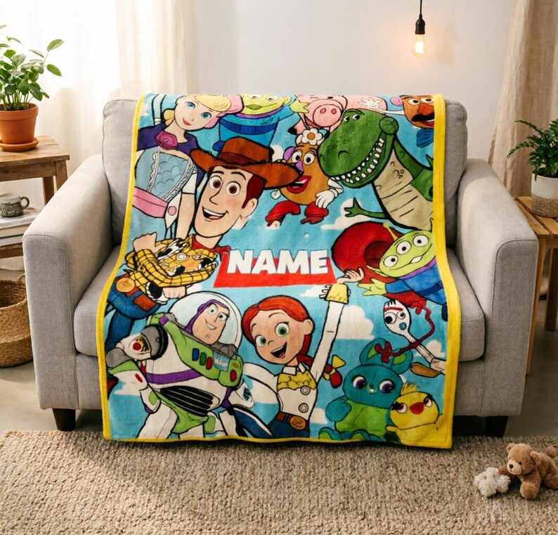 May include: A soft fleece throw blanket featuring a colorful Toy Story character print with Woody, Buzz Lightyear, Jessie, and other figures on a light blue background. The blanket has a yellow border and a central white rectangle with the text NAME.