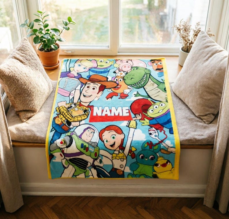 May include: A rectangular fleece blanket featuring a colorful print of various Toy Story characters including Woody, Buzz Lightyear, Jessie, Rex, and the Aliens. The center of the blanket displays a red rectangular graphic with the text NAME.