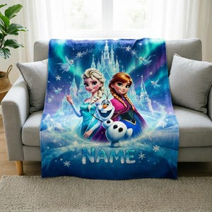 Puede incluir: Una manta rectangular de forro polar con un estampado gr&aacute;fico en azul y morado vibrante de Elsa, Anna y Olaf de Frozen. El dise&ntilde;o incluye un castillo de hielo, copos de nieve y la palabra NAME en letras grandes de color plateado.