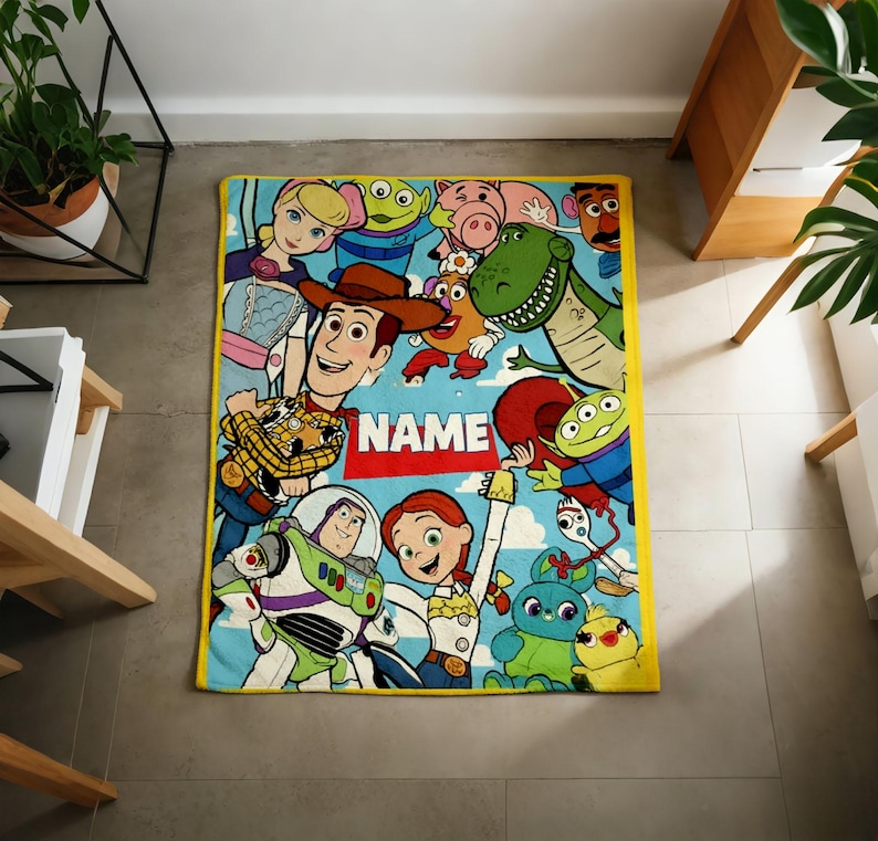 May include: A rectangular fleece blanket with a yellow border featuring a colorful print of various animated characters from the Toy Story franchise, including Woody, Buzz Lightyear, and Jessie, with a red banner in the center displaying the word NAME.