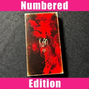 Pode incluir: Uma caixa de cassete retangular com um gráfico em vermelho e preto de um artista no palco. O texto Korn está impresso em branco, com as palavras Wood stock 99 abaixo. O design geral é escuro com efeitos de luz vermelha de alto contraste.