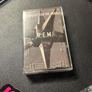 Puede incluir: Una cinta de casete del álbum "Automatic for the People" de R.E.M. La portada presenta un diseño gráfico en blanco y negro con el nombre de la banda y el título del álbum. El casete está en una funda de plástico transparente.