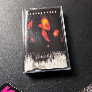 May include: A Soundgarden cassette tape in a clear plastic case. The cover art features a dark, textured background with abstract, glowing orange and red shapes resembling flames or light, and the word SOUNDGARDEN printed in white at the top.