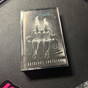 May include: A cassette tape featuring the album art for Jane's Addiction's "Nothing's Shocking." The cover shows a monochrome image of two nude women with flames above their heads, seated on a rocking chair.
