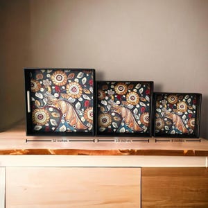 May include: Three square black serving trays with integrated handles, featuring a peacock and floral pattern in shades of gold, blue, and white. The trays are labelled with widths of 30.48 cm, 25.4 cm, and 20.32 cm respectively.