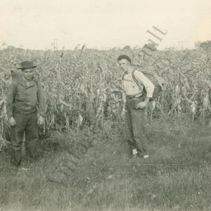 Op de afbeelding: Een zwart-witfoto van twee mensen in een veld met gewassen. Eén persoon draagt een hoed en jas, de ander een rugzak. De scène speelt zich buiten af, met een heldere lucht op de achtergrond.