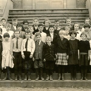 May include: A vintage black and white photograph of a group of children posing on the steps of a building. The children are dressed in formal attire, including dresses, skirts, suits, and ties. The photograph is likely from the early 20th century.