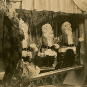 May include: Vintage black and white photograph of a puppet show featuring three Santa Claus figures. The puppets are dressed in red suits with white trim and long white beards. The stage has a decorative railing.