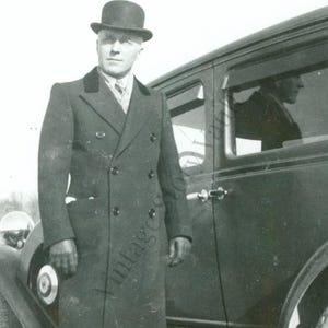 May include: Vintage black and white photograph of a person wearing a dark overcoat and bowler hat, standing next to a classic car. The coat is double-breasted with a contrasting collar. The car has a vintage design.