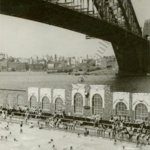 May include: Vintage black and white photograph of a swimming pool with people in the water and sitting on the edge. A large bridge spans the top of the image, with a city skyline in the background.