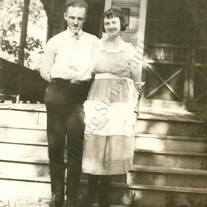 May include: Vintage black and white photograph of a man and a woman standing on the steps of a house. The man wears a collared shirt and dark pants. The woman wears a dress and apron. The photo is likely from the early 20th century.